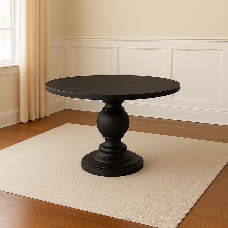 May include: A round, black dining table with a pedestal base. The table has a smooth, dark surface and a sturdy, decorative base. The table is set on a neutral-colored rug in a room with light-colored walls and curtains.
