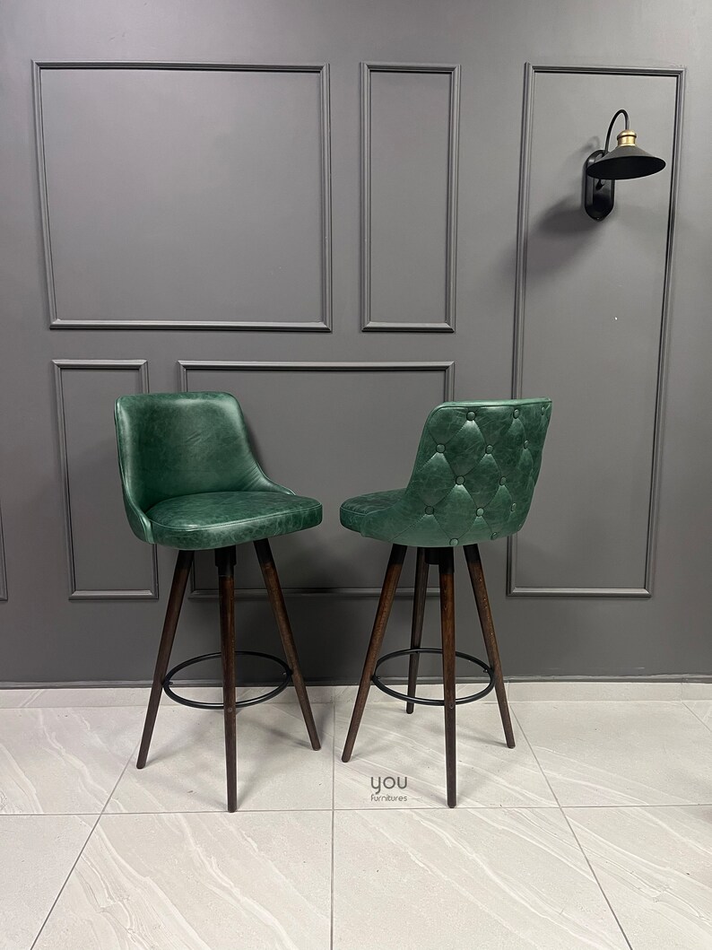 May include: Two green leather bar stools with a tufted design and dark wood legs. The stools have a black metal footrest.