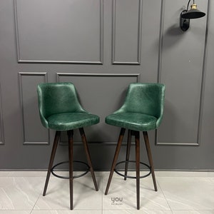 May include: Two green leather bar stools with dark wood legs and a black metal footrest. The stools are set against a grey wall with decorative moulding.