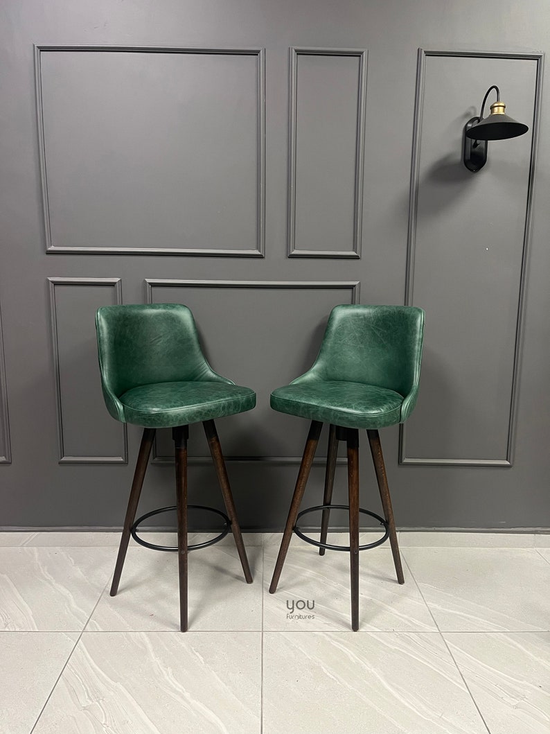 May include: Two green leather bar stools with dark wood legs and a black metal footrest. The stools are set against a grey wall with decorative moulding.
