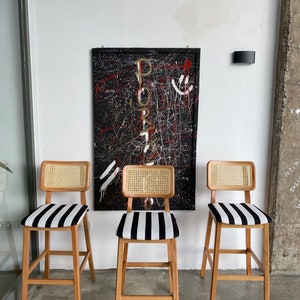 May include: Three wooden bar stools with black and white striped seats and woven cane backs. The stools are in front of a black and white abstract painting with the word "PORTO" written in gold.