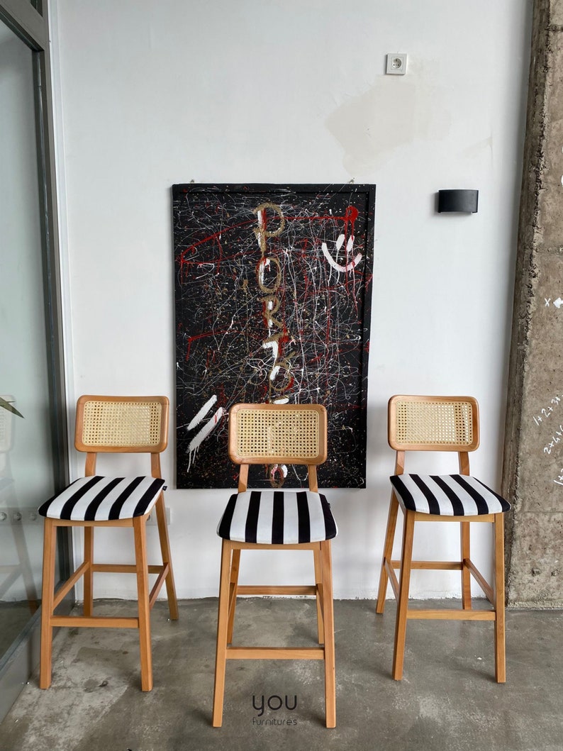 May include: Three wooden bar stools with black and white striped seats and woven cane backs. The stools are in front of a black and white abstract painting with the word "PORTO" written in gold.