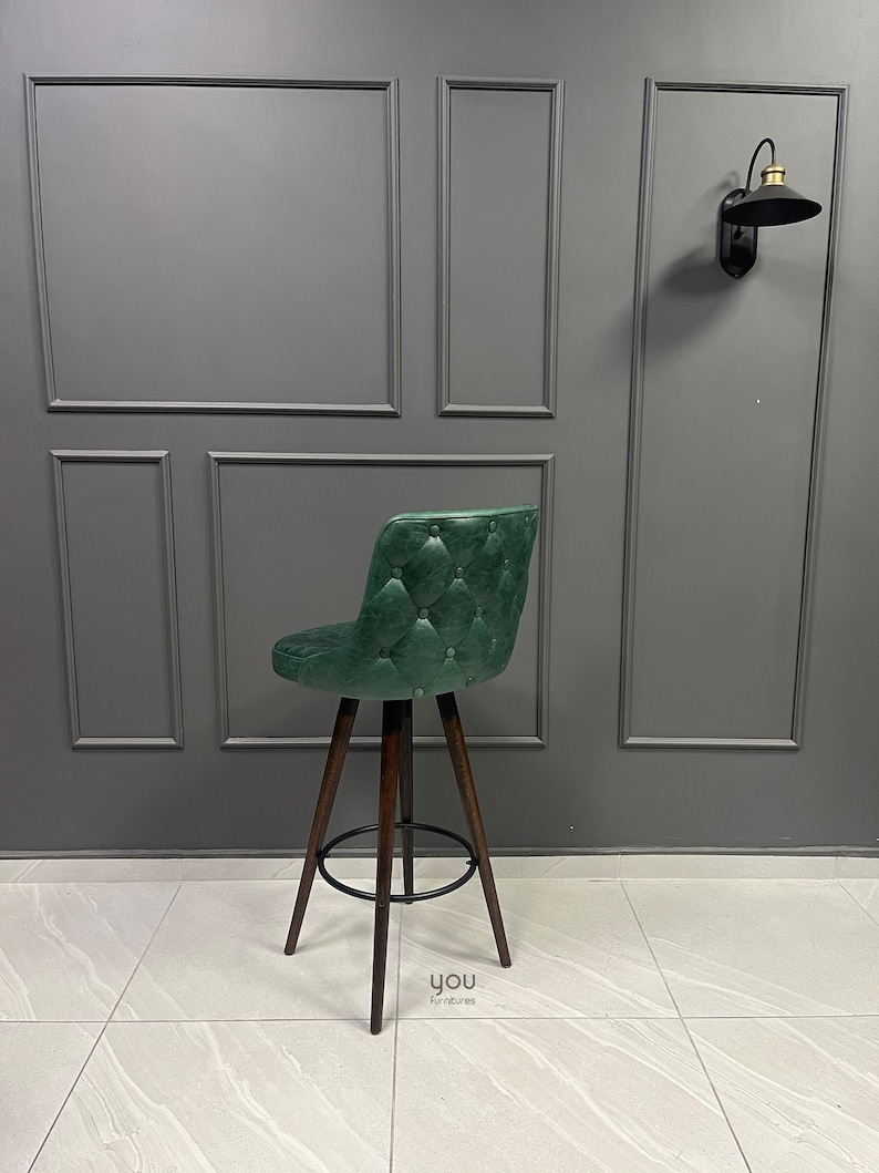 May include: A green upholstered bar stool with a tufted back and wooden legs. The stool is in front of a grey wall with decorative moulding.