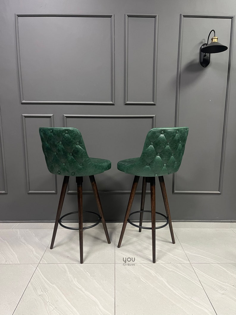May include: Two green upholstered bar stools with a tufted design and dark wood legs. The stools are set against a grey wall with moulding.