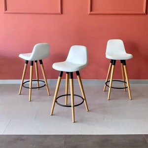 May include: Three white upholstered bar stools with black metal accents and wooden legs. The stools are arranged in a row on a white floor.