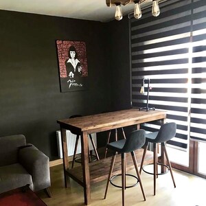 May include: A rustic wooden table with a metal frame. The table has a shelf underneath and is paired with three black bar stools. The table is in a room with a dark grey wall and a window with black blinds.