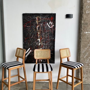 May include: Three wooden bar stools with black and white striped seats and woven cane backs. The stools are in front of a black and white abstract painting.