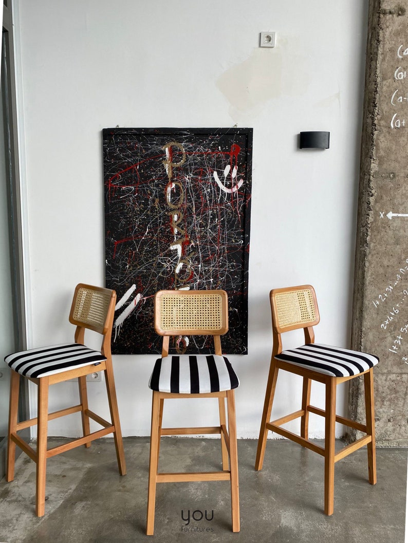 May include: Three wooden bar stools with black and white striped seats and woven cane backs. The stools are in front of a black and white abstract painting.