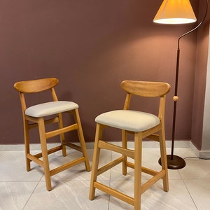 May include: Two wooden bar stools with light beige fabric seats. The stools have a simple, modern design with a curved backrest and a single crossbar for support. The stools are set against a brown wall with a floor lamp in the background.