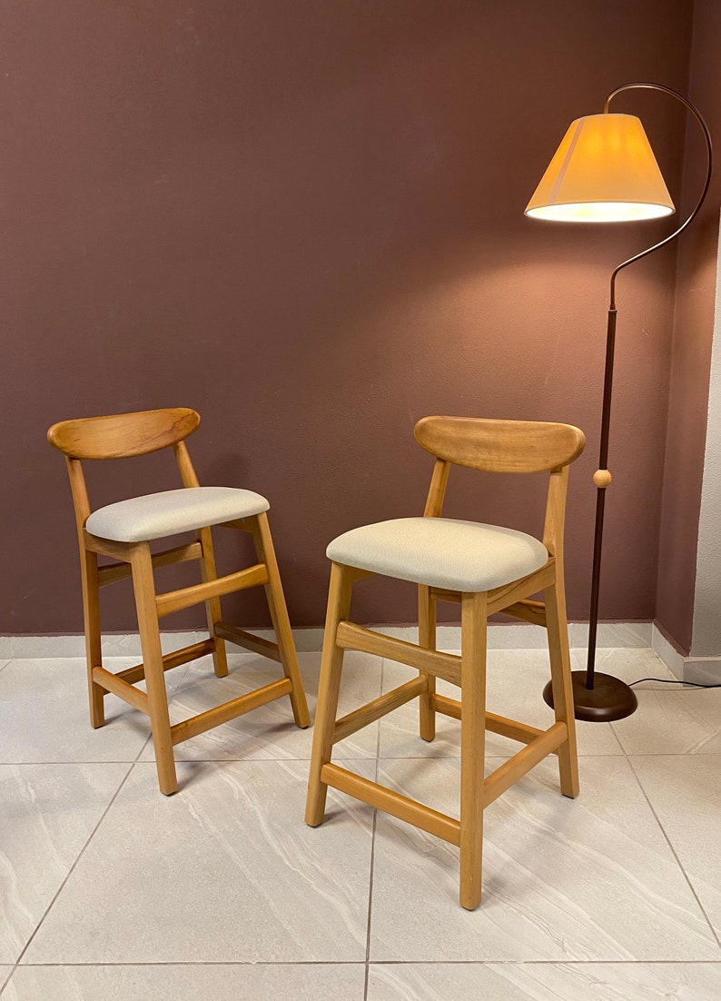 May include: Two wooden bar stools with light beige fabric seats. The stools have a simple, modern design with a curved backrest and a single crossbar for support. The stools are set against a brown wall with a floor lamp in the background.