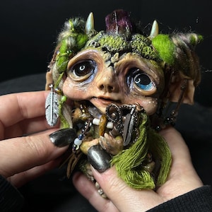 Puede incluir: Escultura detallada de una criatura fantástica con ojos grandes, adornada con musgo verde, plumas y un cristal púrpura. La figura sostiene un objeto pequeño y está adornada con cuentas y otros elementos decorativos. La paleta de colores incluye tonos tierra y verdes vibrantes.