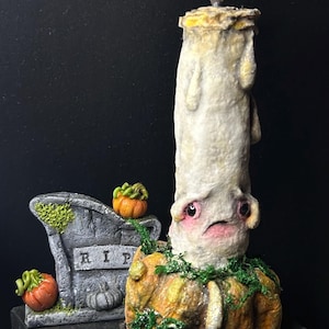 Op de afbeelding: Een grillige Halloween-kaars sculptuur. De kaars is wit met een gezicht en staat op een pompoenbasis. Een kleine grafsteen met "RIP" staat naast de kaars, samen met miniatuurpompoenen. De kaars heeft een brandende vlam.