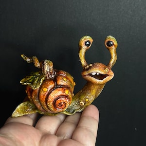 Könnte beinhalten: Eine kleine, handbemalte Skulptur einer Schnecke mit einem kürbisförmigen Gehäuse. Das Wesen hat einen strukturierten, orange-grün gesprenkelten Körper, zwei markante Augenstiele mit schwarzen Pupillen und ein weit geöffnetes Maul mit Zähnen.