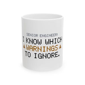 May include: A white ceramic mug featuring pixelated black and brown text that reads: SENIOR ENGINEER: I KNOW WHICH WARNINGS TO IGNORE. The design includes two small brown pixelated traffic cone icons flanking the word WARNINGS.