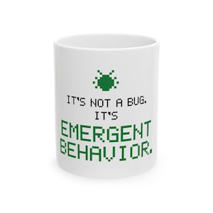 May include: A white ceramic mug featuring a green pixelated insect icon at the top. Below the icon, black and green pixelated text reads: IT'S NOT A BUG. IT'S EMERGENT BEHAVIOR. The mug has a cylindrical shape and a smooth, glossy finish.