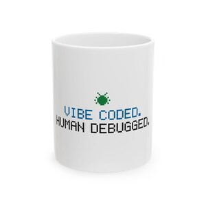 May include: A white ceramic mug featuring a green pixelated bug icon centered above the blue pixelated text VIBE CODED. and the black pixelated text HUMAN DEBUGGED. The design is printed on the front of the smooth, cylindrical vessel.