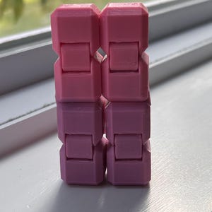 May include: A pink and purple fidget toy, composed of interlocking rectangular blocks. The toy is made of plastic and has a smooth texture. The color gradient transitions from a light pink to a darker purple. The toy is designed to be manipulated.