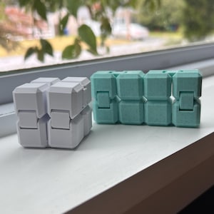 May include: Two fidget cubes, one white and one teal, sit side-by-side on a white surface. Each cube is composed of multiple interlocking blocks, allowing for movement and manipulation. The background shows a blurred view of greenery and a window.