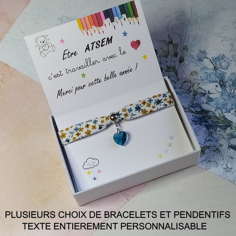 May include: A white gift box with a blue and yellow patterned bracelet inside. The box has a card with the text "Etre ATSEM c'est travailler avec le Merci pour cette belle ann&eacute;e!" and a drawing of a teddy bear. The box also has a cloud with raindrops and stars. The bracelet has a blue heart charm.
