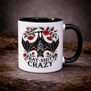 11oz ceramic two-tone mug featuring a detailed bat illustration with red floral elements and “Bat Shit Crazy” text. Styled on a textured stone background with a dark, moody aesthetic. Ideal for gothic gifts, dark humour lovers, and alternative homeware.