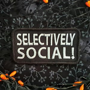 May include: A black patch embroidered with the words "SELECTIVELY SOCIAL!" in white. The patch is set against a dark floral background, with a garland of orange and black decorations.