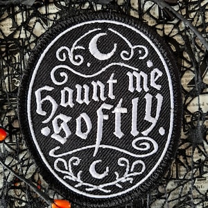 May include: A circular black patch embroidered with the words "Haunt me softly" in white. The design includes crescent moons and decorative swirls. The patch is set against black netting and orange beads.