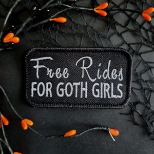 May include: A black rectangular patch with the words "Free Rides FOR GOTH GIRLS" embroidered in white script. The patch is set against a black background with orange and black decorative elements.