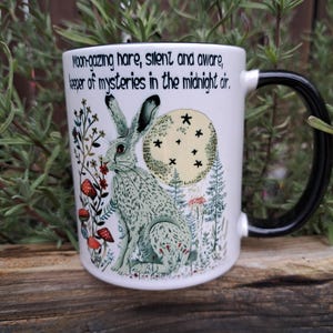 May include: White ceramic mug with a black handle. The mug features a colourful illustration of a hare sitting in a forest with a full moon behind it. The text on the mug reads: "Moon-gazing hare, silent and aware, keeper of mysteries in the midnight air."