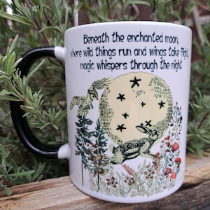 A stunning 11oz ceramic coffee mug with a whimsical moon-gazing hare under a starry night sky, surrounded by lush forest flora and a soaring owl. The quote, "Beneath the enchanted moon, where wild things run and wings take flight, magic whispers through the night," enhances its mystical charm. Perfect for witchy decor and nature lovers.