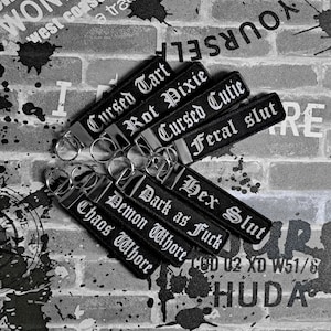 May include: Black keychains with silver key rings. Each keychain has white gothic lettering on a black background. The phrases include "Cursed Tart", "Rot Pixie", "Cursed Cutie", "Feral Slut", "Hex Slut", "Dark as Fuck", "Demon Whore", and "Chaos Whore".