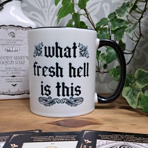 May include: White ceramic mug with a black rim and handle. The mug has black gothic lettering that reads "what fresh hell is this".