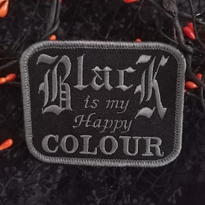 May include: A black rectangular patch with grey embroidered text. The text reads "Black is my Happy COLOUR". The patch has a grey border and is set against a dark background with floral patterns and orange accents.