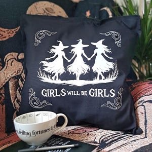 May include: Black throw pillow with a white graphic of three witches holding hands, with the text "GIRLS WILL BE GIRLS". A white teacup with the text "when telling fortunes in tea" is also visible.