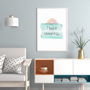 May include: A framed print with a watercolor background in shades of blue and green. The print features a simple line drawing of a sun setting over the ocean with the text "Make Waves" in black.