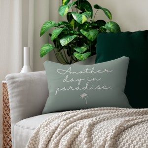 May include: A light green throw pillow with white text that reads "Another day in paradise."  A small palm tree graphic is below the text. The pillow is on a light grey sofa with a white throw blanket.