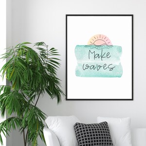May include: A framed print with a watercolor background in shades of blue and green. The print features a stylized sun with a pink and orange gradient and the text "Make waves" in black.