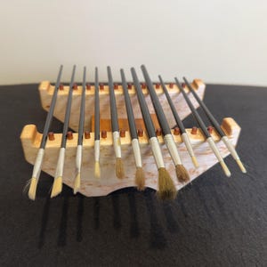 May include: A collection of paint brushes with dark gray handles and white bristles, displayed in a wooden brush holder. The brushes vary in size and bristle shape, suitable for various painting techniques.
