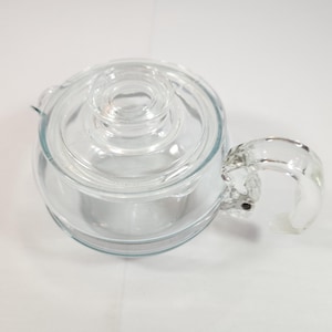 May include: A clear glass teapot with a rounded body and a matching lid. The teapot has a curved handle and a spout. The design is simple and functional, suitable for brewing and serving tea.
