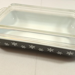 May include: Rectangular baking dish with a clear glass lid. The dish is dark gray with white snowflake designs along the sides. The lid sits on top of the dish. A vintage kitchenware item.