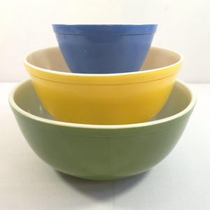 May include: A stack of three colorful mixing bowls. The largest bowl is green, the middle bowl is yellow, and the smallest bowl is blue. Each bowl has a white interior rim.