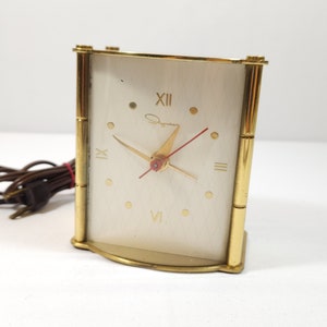 May include: A gold-toned electric alarm clock with a white face and black Roman numeral hour markers. The clock has a red second hand and a black minute hand. The clock is made by the company "Symphony".