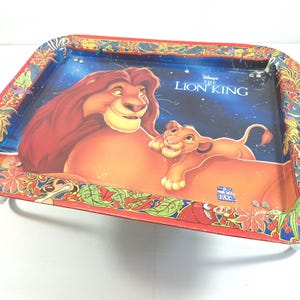 May include: A rectangular metal tray featuring a colourful illustration of Simba and Mufasa from The Lion King. The border is decorated with a vibrant jungle leaf pattern in red, green, and yellow. The text The Lion King is printed in white.