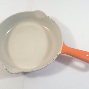 May include: A round enamelled cast iron skillet with a cream-coloured interior and a bright orange exterior. The pan features a single handle with a hole for hanging and two small pouring spouts on opposite sides of the rim, showing minor wear.