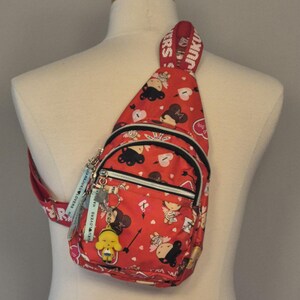 May include: A red sling backpack featuring a repeating pattern of cartoon characters, hearts, and arrows. The bag has a front zippered pocket, a main compartment with a zipper, and a wide shoulder strap with the text JUKU LOVERS printed in white.