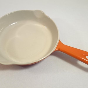 Peut inclure: Une poêle avec un manche orange et un intérieur crème. La poêle a une forme arrondie avec un bec verseur et un trou au bout du manche. L'ustensile de cuisine est conçu pour une utilisation sur la cuisinière.