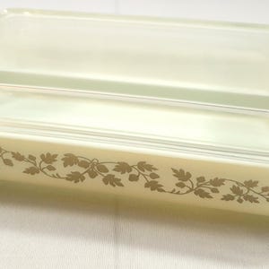 Könnte beinhalten: Eine rechteckige, cremefarbene Pyrex-Auflaufform mit einem Deckel aus klarem Glas. Die Form hat ein dekoratives goldenes Blattmuster an der Seite. Der Deckel liegt bündig auf der Form, die Griffe hat.