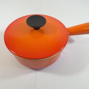 May include: A round, orange enameled cast iron saucepan with a matching lid and a single long handle. The lid features a black circular knob in the center. The exterior of the pan transitions from a bright orange to a deeper red-orange hue.