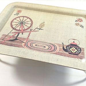 May include: A vintage serving tray with a beige linen-effect background. The tray features a spinning wheel, a teapot, and floral designs in pink, brown, and black. The tray has clear plastic legs.