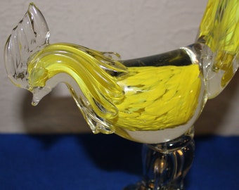Könnte beinhalten: Eine gelbe Glas-Hahn-Figur mit klaren Glasdetails. Der Hahn hat einen langen, geschwungenen Hals und einen großen, detaillierten Kamm. Der Sockel der Figur ist aus klarem Glas.