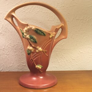 May include: A pink and green ceramic vase with a floral design. The vase has a wide, open mouth and a narrow base. It is decorated with a pattern of stylized flowers and leaves.